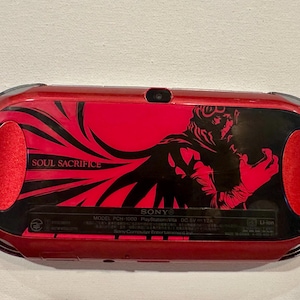 May include: A red and black Sony PlayStation Vita handheld gaming console featuring a Soul Sacrifice graphic on the back panel. The device includes a central black section with the text Soul Sacrifice and a character illustration, flanked by red speakers.