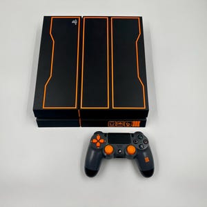 May include: A black video game console featuring orange rectangular outlines and a silver PlayStation logo. The front panel displays three orange icons and the Roman numeral III. A matching black and gray wireless controller with orange buttons.