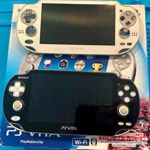 May include: A silver and a black Sony PS Vita handheld gaming console resting on a blue and white product box. The devices feature a central screen, directional pad, action buttons, and dual analog sticks. The box displays the PS Vita logo and text.