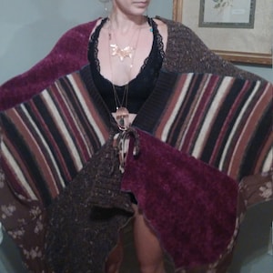 May include: A brown, burgundy, and black patchwork knit poncho with fringe detail. The poncho is worn over a black lace bralette and layered necklaces.