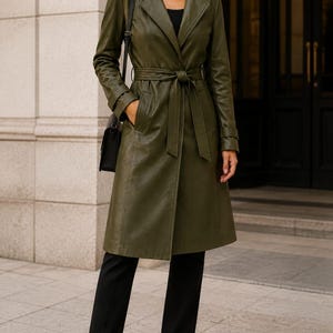 Könnte beinhalten: Ein olivgrüner Trenchcoat mit Reverskragen, langen Ärmeln und einem passenden Bindegürtel in der Taille. Der Mantel hat eine glatte Oberfläche, Seitentaschen und geknöpfte Ärmelabschlüsse. Er wird über einem schwarzen Oberteil und einer schwarzen Hose getragen.