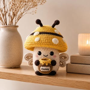 May include: A crocheted mushroom figure with a yellow cap featuring white spots and a dark brown band. The cream-colored body has black eyes, a small smile, pink cheeks, and small wings. It holds a brown honey pot with the word Honey printed on it.
