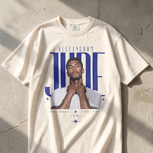 Könnte beinhalten: Ein cremefarbenes Kurzarm-T-Shirt mit einem Grafikdruck von Jude Bellingham. Das Design enthält den Text BELLINGHAM JUDE, REAL MADRID, ENGLAND und die Zahl 10, ergänzt durch kleine Sternsymbole und ein zentrales Porträt des Sportlers.