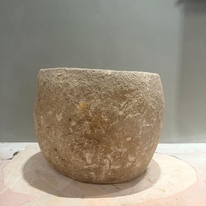 May include: A large, cylindrical stone vessel with a rough, weathered texture and a natural beige and tan color palette. The object features a wide, open top and a solid, thick-walled construction with visible surface irregularities and earthy tones.