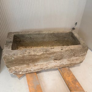May include: A large, rectangular stone trough with a rough, weathered exterior and a hollowed-out center. The natural beige and grey stone features a porous, uneven texture throughout its surface, showing signs of age and natural wear.