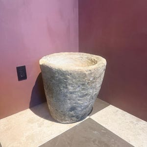 May include: A large, heavy, cylindrical stone vessel with a rough, weathered exterior texture and a wide, hollowed-out opening at the top. The stone displays natural variations in color, ranging from light beige to grey, with a porous surface.
