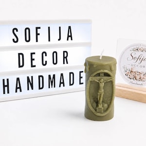 Könnte beinhalten: Eine zylindrische olivgrüne Kerze mit einem erhabenen Relief eines Kruzifixes in einem ovalen Rahmen. Daneben steht eine weiße Leuchtbox mit dem schwarzen Text SOFIJA DECOR HANDMADE sowie ein rundes Schild mit dem Logo von Sofija Decor.