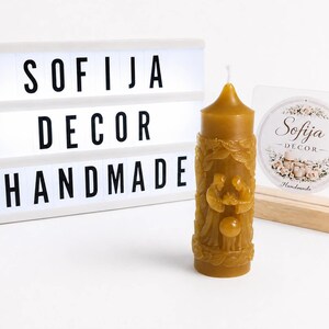 Könnte beinhalten: Eine zylindrische, goldene Bienenwachskerze mit einem Relief einer Krippenszene, neben einer weißen Leuchtbox mit dem schwarzen Text SOFIJA DECOR HANDMADE und einem kleinen transparenten Schild mit einem runden Logo und floralen Details.