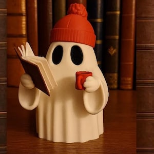 May include: A white ghost figurine wearing a bright orange beanie with a pom-pom. The ghost has large black oval eyes and is holding an open brown book in one hand and a small red mug in the other, standing against a backdrop of old books.