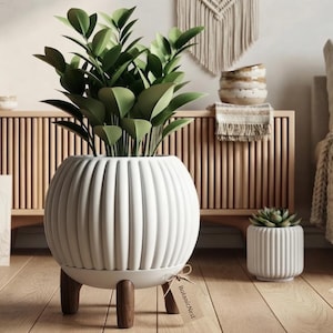Op de afbeelding: Een grote witte, geribbelde, bolvormige plantenbak met drie donkerbruine houten poten, met daarin een groene plant. Een klein label met de tekst BotanicNest is aan de basis bevestigd. Een kleinere, bijpassende witte plantenbak staat rechts.