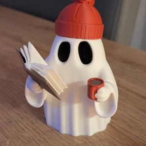 Op de afbeelding: Een wit spookfiguurtje met een feloranje muts met een pompon bovenop. Het spook heeft grote zwarte ovale ogen, houdt een open boek in de ene hand en een kleine oranje mok gevuld met een donkerbruine vloeistof in de andere hand.