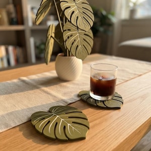May include: Two olive green coasters shaped like monstera leaves with white vein detailing. One coaster sits flat on a wooden surface, while the other supports a clear glass filled with a dark brown liquid and ice cubes, reflecting a natural aesthetic.