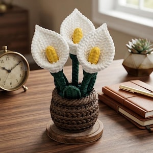 Pode incluir: Um arranjo decorativo de três copos-de-leite em crochê com pétalas brancas e espádices amarelos, sustentados por caules verde-escuros. As flores estão contidas num vaso castanho de textura entrançada sobre uma base circular de madeira.
