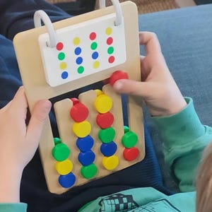 May include: A wooden board game featuring four vertical tracks with sliding circular tokens in red, yellow, blue, and green. A white rectangular header at the top displays a matching color pattern of dots in four rows and four columns.