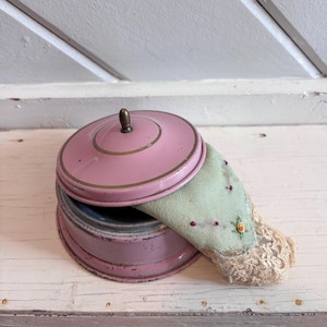 May include: A round, pink metal container with a matching lid and a small brass-colored knob. A light green fabric pincushion with delicate floral embroidery and beige lace trim is tucked inside the container, partially resting on the lid.