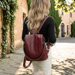 May include: A deep burgundy backpack with a smooth finish, featuring vertical stitched detailing, a top handle, and adjustable shoulder straps. The bag has a structured silhouette with visible metal hardware attachments on the sides and top.