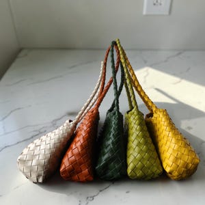 Puede incluir: Cinco bolsos pequeños tejidos dispuestos en fila, cada uno con un asa trenzada. Los bolsos son de color crema, naranja quemado, verde oscuro, verde lima y amarillo brillante, todos con una textura tejida de patrón cuadrado.
