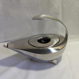 May include: A polished stainless steel teapot with a teardrop-shaped body, a single curved handle extending over the top, a small spout, and a circular lid featuring a black knob. The vessel is supported by three small, integrated metal feet.