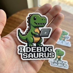 Puede incluir: Una pegatina de vinilo troquelada que muestra un dinosaurio verde de arte píxel con una barriga color canela, sosteniendo un portátil gris. El texto debajo del dinosaurio dice I DEBUG SAURUS en una fuente negra, negrita y pixelada.