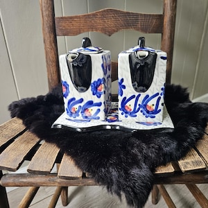 May include: Two square ceramic containers with lids and a matching rectangular tray. The white ceramic features a hand-painted pattern of blue flowers with red centers and black accents. Each container has a black spout and a small lid on top.