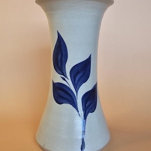 May include: A tall, flared ceramic vase with a speckled off-white finish. The body of the vase is decorated with a hand-painted dark blue botanical motif featuring a central stem with several pointed leaves extending outward in a vertical pattern.