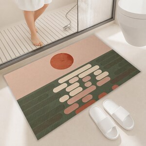 May include: A rectangular bath mat featuring a graphic design with a solid terracotta sun against a light pink sky above a dark green landscape. The lower section displays a pattern of horizontal, rounded bars in shades of cream, pink, and terracotta.
