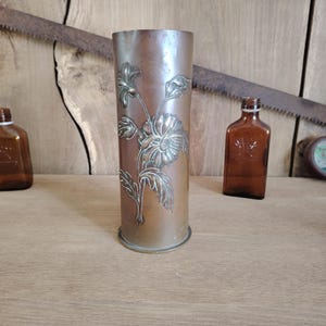 May include: A cylindrical brass shell casing vase featuring an embossed floral design of a large central flower with leaves and stems. The metal has a weathered, aged patina with a circular base and an open top, standing upright as a decorative vessel.