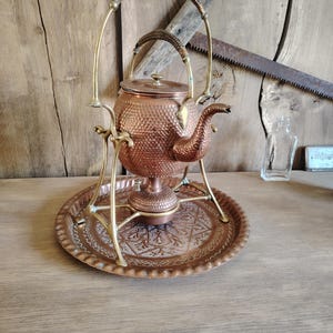 Peut inclure: Une bouilloire en cuivre martelé avec une anse et un bec en laiton, suspendue dans un support en laiton au-dessus d'un petit brûleur en cuivre. L'ensemble repose sur un plateau rond en cuivre gaufré à bord festonné et motifs floraux.