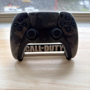 May include: A gray and black camouflage patterned video game controller featuring a central PlayStation logo and two analog sticks. The controller rests on a black stand with the white text CALL OF DUTY printed across the front base.