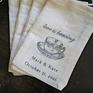 May include: Set of six small, off-white fabric bags with a drawstring closure. Each bag has a black and white illustration of a teacup and the text "love is brewing" and "Mark & Kaite October 31, 2016".