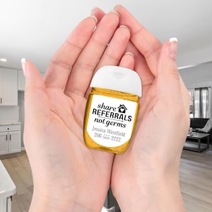 A small, clear, yellow hand sanitizer bottle with a white cap is cradled in two hands. The label says "share referrals not germs" with a house icon and a line through the word "germs". The label also includes a name and phone number: "Jessica Westfield 206-555-2222".