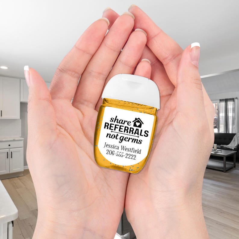 A small, clear, yellow hand sanitizer bottle with a white cap is cradled in two hands. The label says "share referrals not germs" with a house icon and a line through the word "germs". The label also includes a name and phone number: "Jessica Westfield 206-555-2222".