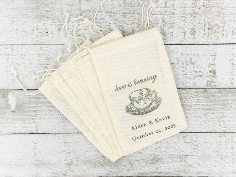 May include: A set of ten natural-colored muslin bags with a drawstring closure. The bags are printed with the text "love is brewing" and a vintage-style illustration of a teacup and saucer. The text "Alden & Kevin October 22, 2025" is printed below the illustration.