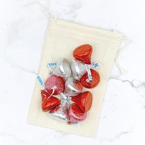Op de afbeelding: Een klein, naturelkleurig stoffen zakje gevuld met rode en roze in folie verpakte chocolade kusjes. Het zakje heeft een trekkoord sluiting en ligt op een wit marmeren oppervlak.