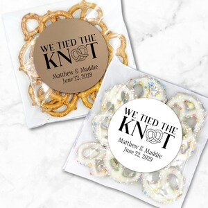 One clear favor bag filled with pretzels has a round brown label attached.  One clear favor bag filled with candy covered pretzels has a white label attached.  Label has text 'We tied the KNOT' with the O replaced by a cute pretzel graphic.  Labels are personalized below the design with names and wedding date.