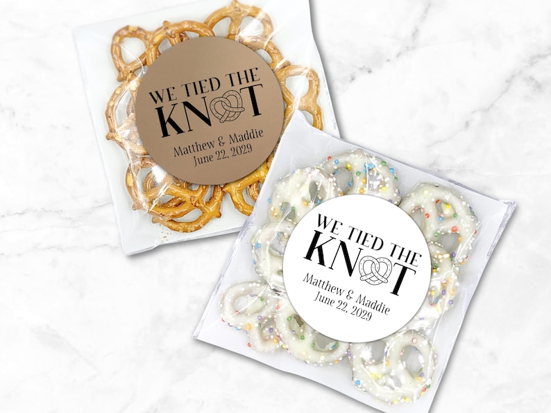 One clear favor bag filled with pretzels has a round brown label attached.  One clear favor bag filled with candy covered pretzels has a white label attached.  Label has text 'We tied the KNOT' with the O replaced by a cute pretzel graphic.  Labels are personalized below the design with names and wedding date.