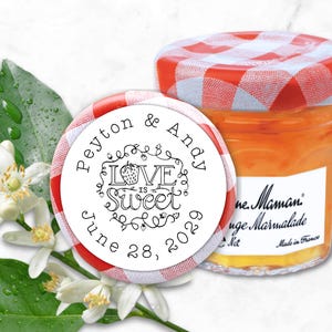May include: A jar of orange marmalade with a red and white checkered lid, next to a round sticker that reads "Love is Sweet" with the names Peyton & Andy and the date June 28, 2022. White flowers and green leaves are in the background.