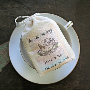 May include: A white fabric bag with a teacup design and the text "love is brewing" on it. The bag is sitting on a white saucer with a gold rim. The text "Mark & Kate October 31, 2016" is printed on the bag.