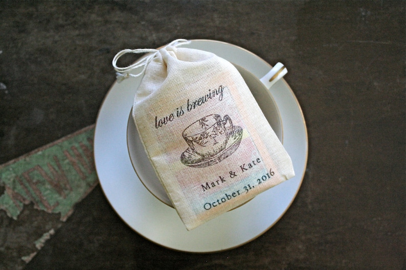 May include: A white fabric bag with a teacup design and the text "love is brewing" on it. The bag is sitting on a white saucer with a gold rim. The text "Mark & Kate October 31, 2016" is printed on the bag.