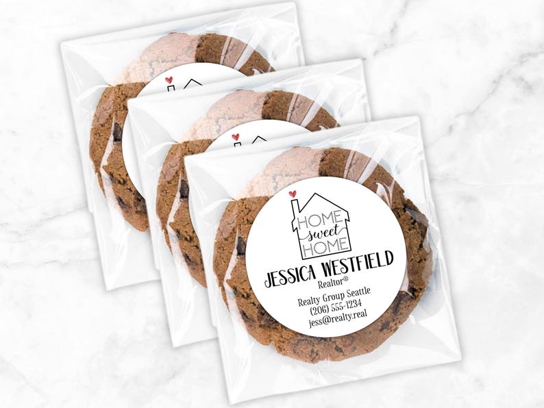 Three clear, square treat bags are shown with chocolate chip cookies inside.  Bags have a round white label attached to the front.  Label design: 'Home Sweet Home' inside a simple house outline, with a small red heart coming out of the chimney.  Labels are personalized at the bottom with professional contact info 'Jessica Westfield, Realtor / Realty Group Seattle / 206-555-1234 / jess@realty.real'