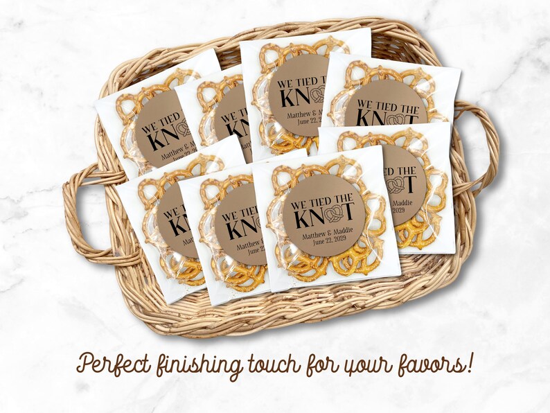 A basket is filled with favor bags.  Each clear bag is filled with brown pretzels and has a kraft paper label attached to the front.  One clear favor bag filled with pretzels has a round brown label attached.  Label has text 'We tied the KNOT' with the O replaced by a cute pretzel graphic.  Labels are personalized below the design with names and wedding date.  Overlay text: 'Perfect finishing touch for your favors!'