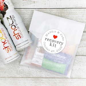 May include: A white paper bag with a round sticker that says "Maddie & Jake recovery kit October 27, 2028" with a red heart. The bag contains various items, including two energy drink bottles, a packet of pills, and a packet of lemon lime seltzer.