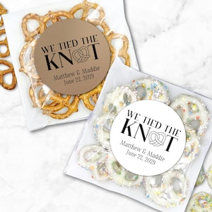 One clear favor bag filled with pretzels has a round brown label attached.  One clear favor bag filled with candy covered pretzels has a white label attached.  Label has text 'We tied the KNOT' with the O replaced by a cute pretzel graphic.  Labels are personalized below the design with names and wedding date.