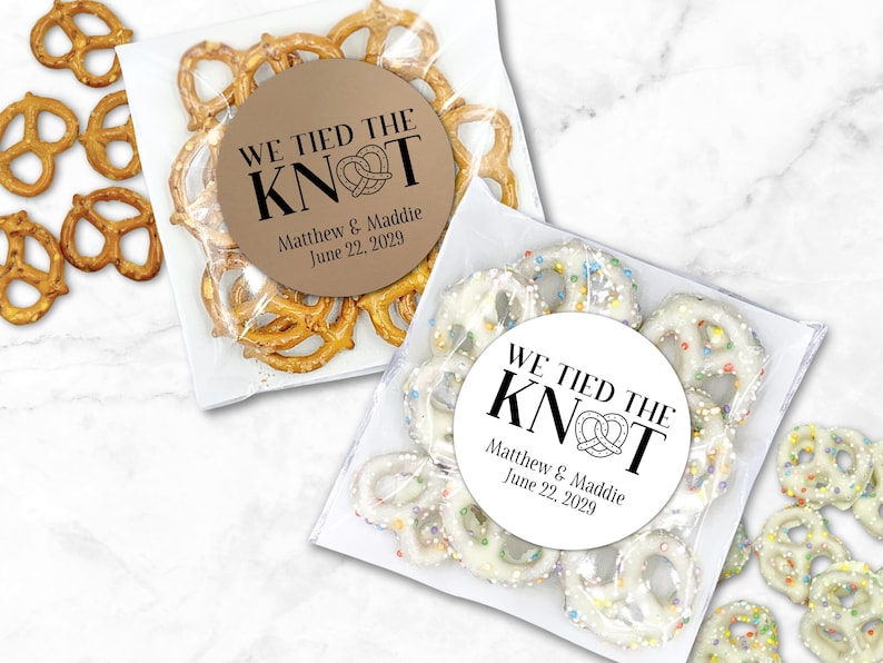 One clear favor bag filled with pretzels has a round brown label attached.  One clear favor bag filled with candy covered pretzels has a white label attached.  Label has text 'We tied the KNOT' with the O replaced by a cute pretzel graphic.  Labels are personalized below the design with names and wedding date.