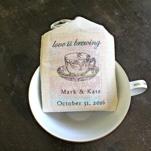 May include: A white tea bag favor with the text "love is brewing" and a vintage illustration of a teacup. The bag also includes the names "Mark & Kate" and the date "October 31, 2016".