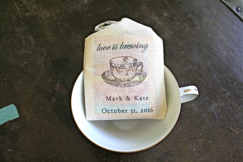 May include: A white tea bag favor with the text "love is brewing" and a vintage illustration of a teacup. The bag also includes the names "Mark & Kate" and the date "October 31, 2016".