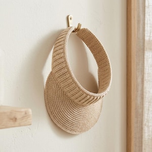 May include: A beige sun visor crafted from a woven material, featuring a wide, rounded brim and a structured, ribbed headband. The visor is suspended from a simple metal hook, showcasing its circular shape and textured, natural-toned construction.