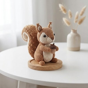 May include: A crocheted squirrel figurine made of brown and cream yarn, featuring a large bushy tail, small pointed ears, and black bead eyes. The squirrel is holding a small brown acorn in its paws and is mounted on a round wooden base.