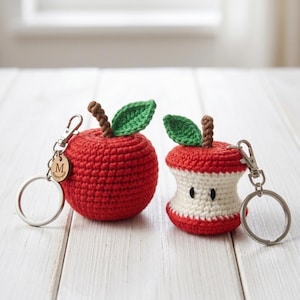 May include: Two crocheted apple keychains. One is a round, solid red apple with a brown stem and green leaf. The other is a red and white apple core with a brown stem and green leaf. Both feature silver metal lobster clasps and key rings.