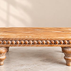 May include: A rectangular wooden riser with a light brown, distressed finish. The platform features a decorative border of small, rounded wooden beads along the edge. The piece is supported by four thick, carved, turned-style legs.